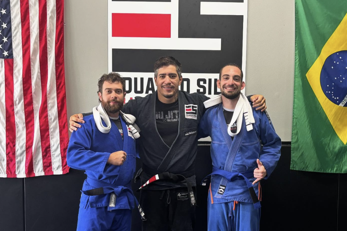 Third-degree black belt Eduardo Silva standing with two Brazilian Jiu-Jitsu students in blue gis at his Texas BJJ and MMA gym, with American and Brazilian flags on the wall
