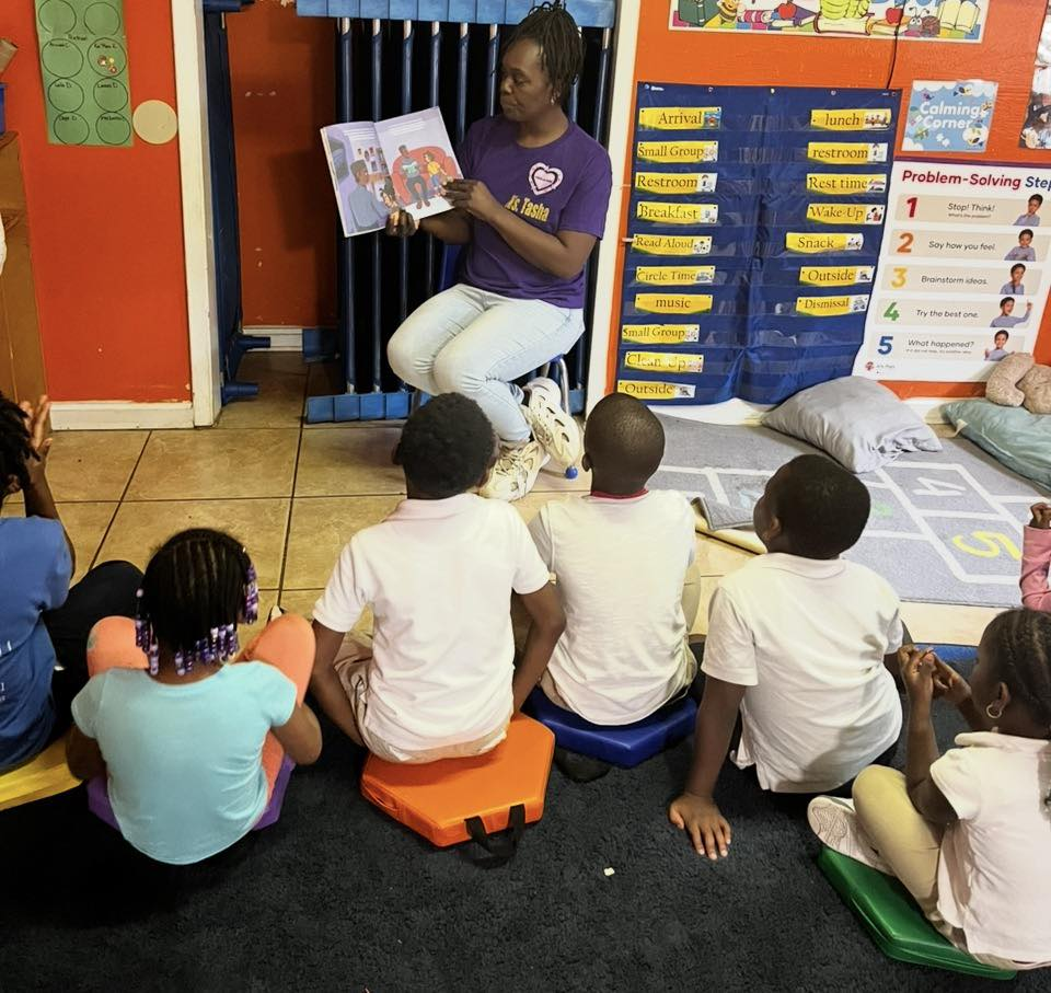 A teacher in a purple shirt reading a picture book aloud to young children seated on colorful cushions in a bright classroom