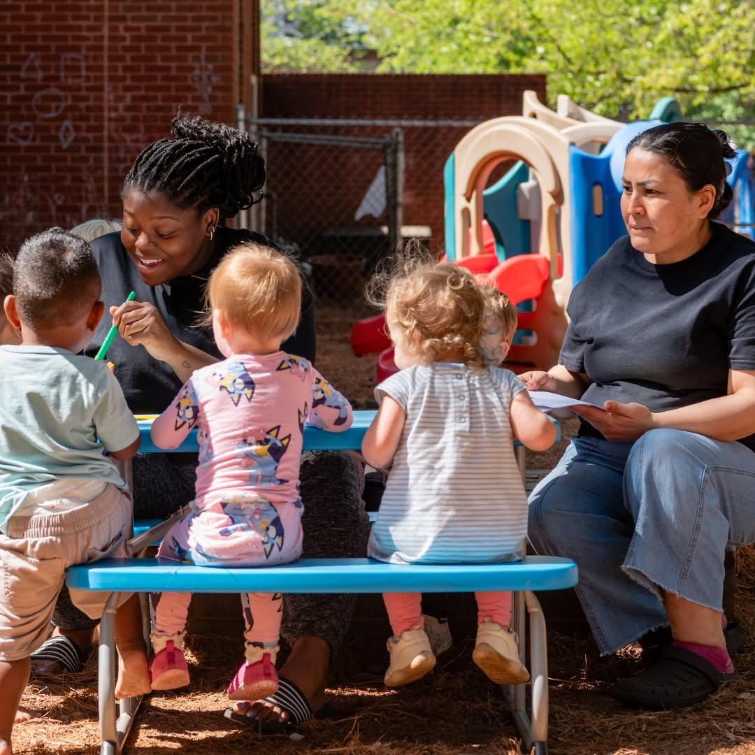 Two teachers in black shirts sitting with three toddlers at a small blue picnic table outdoors, drawing with chalk on a brick wall beside a playground